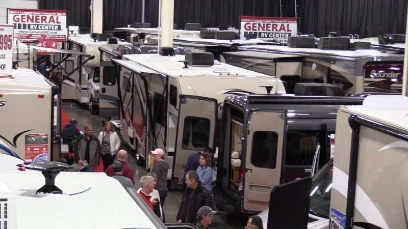 Many RVs are setup with doors open and on display inside a convention center during an RV show. Attendees are walking in the aisle and looking at the different RVs.