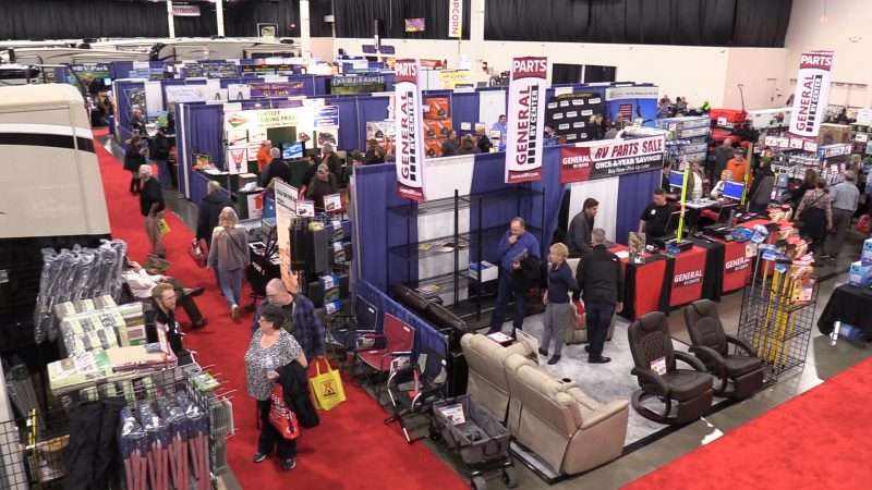 Several men and women walk the aisles of a large convention center where booths filled with RV accessories are on display. The booth in the foreground shows RV chairs and other accessories. There are RVs visible on the far left and in the background. 