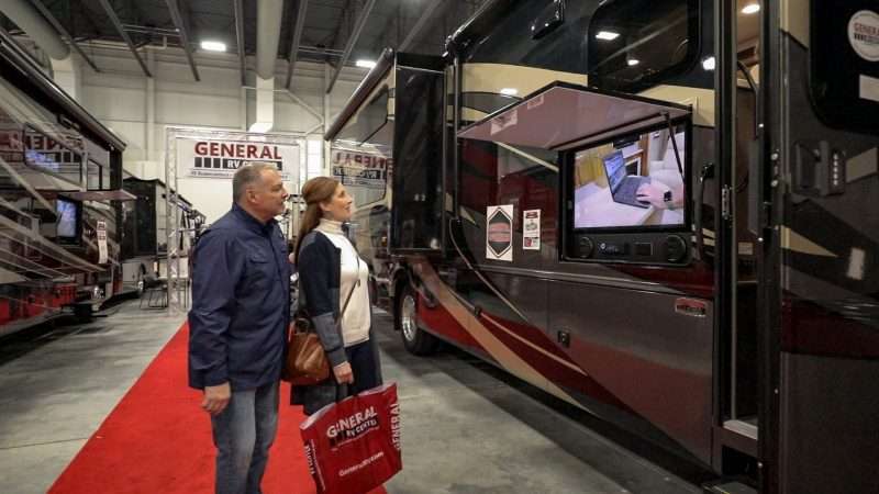 A man and woman are watching a video that's playing on a TV mounted to the exterior of a large RV. The RV is set up inside a convention center for an RV show. 