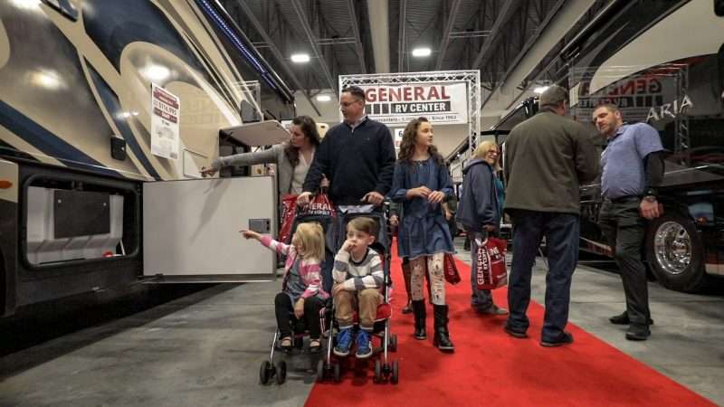 A family with a mother, father, older daughter, and two younger children attend an RV show. The youngest boy and girl and sitting in a stroller and being pushed by their father. The mother and older daughter are looking at display RVs on either side of the aisle. They are walking past another couple speaking with a manufacturer representative.