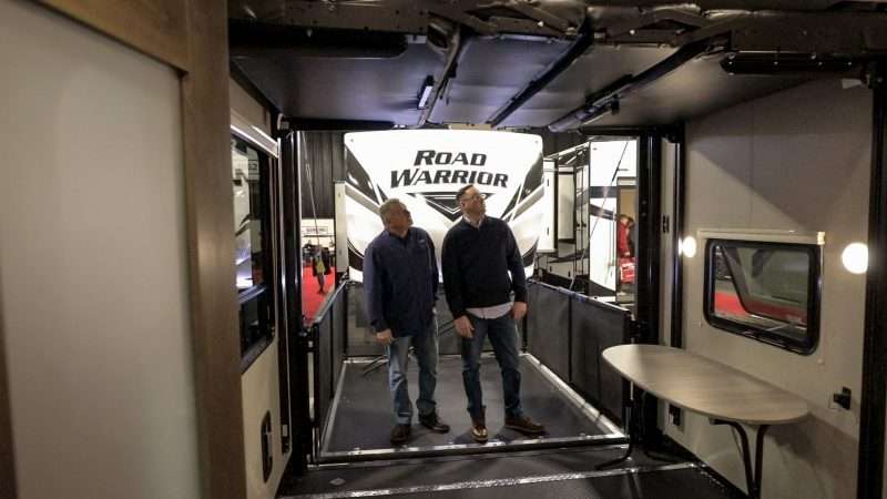 Two men stand inside an RV on display at an RV show. They are looking at two retractable bunks that can be lowered from the ceiling and double as beds. The RV they are looking at also has a fold-down rear door that doubles as a deck patio, complete with railing.  