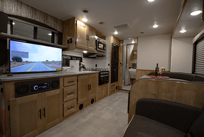 A photo of Coachmen RV's Leprechaun 319M RV interior shows off it's modern decor. Light, cherry colored cabinets with dark hardware, dark fabrics, and black appliances. 