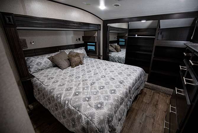 A photo of the RV master bedroom of the Jayco Eagle 317RLOK shows the modern decor: light fabrics and light gray walls contrast with dark wood cabinets.