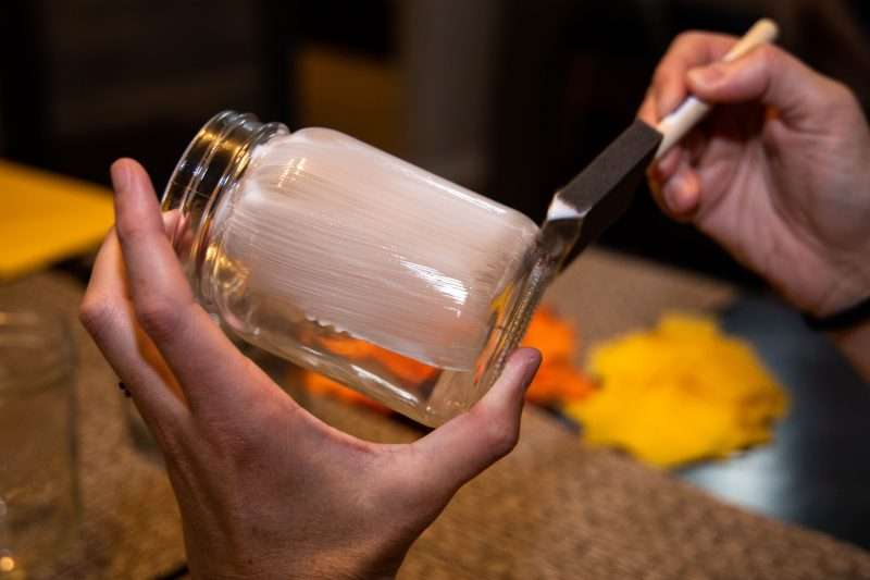 A woman spreads glue on a mason jar with a sponge brush.