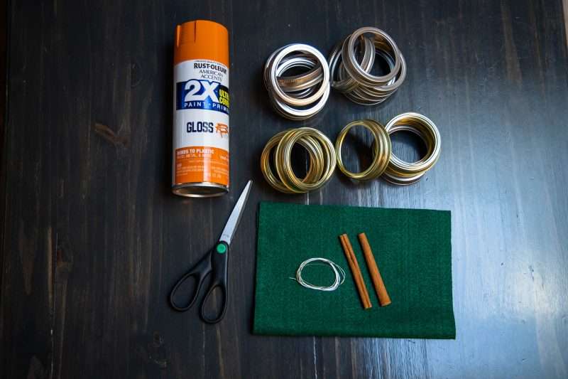 A can of orange spray paint, scissors, green felt, wire, two cinnamon sticks and mason jar lid rings are arranged on a wooden background.