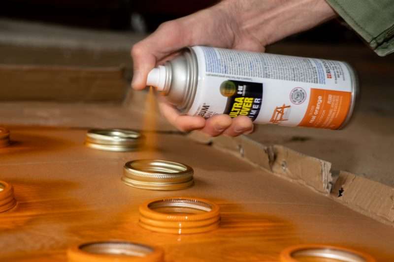 A woman spray paints mason jar lids with orange spray paint.