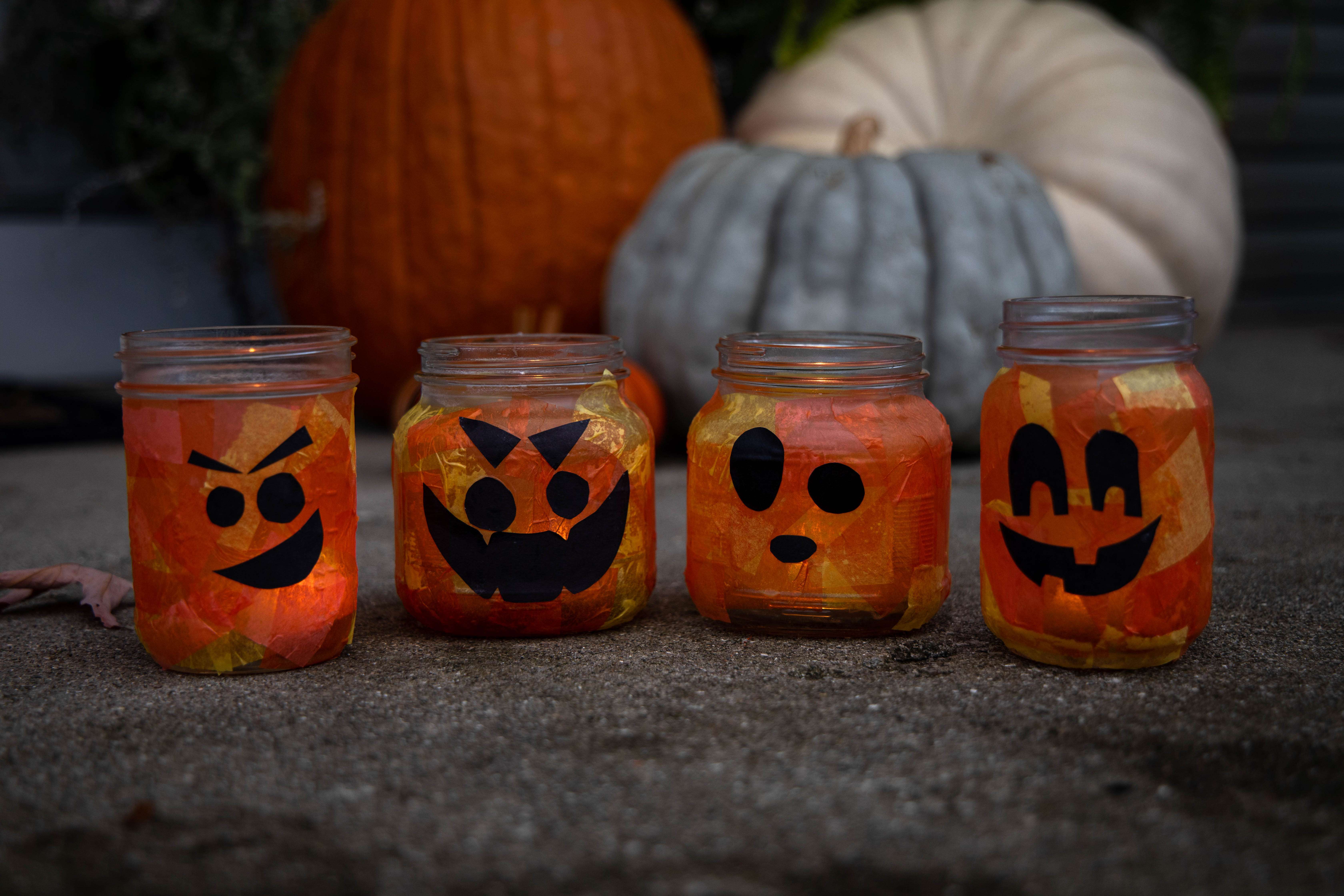 Mason jar pumpkins sit on the ground in front of real pumpkins and squash for a Halloween-themed scene.