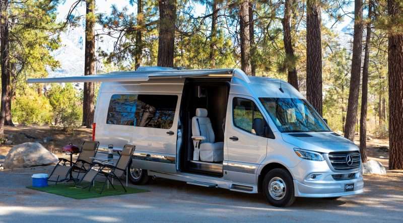 A silver Grech Strada Class B RV is parked at a wooded campsite with its awning extended. Below the awning, there are two lounge chairs, a grill, cooler, and folding table with a lantern set up.