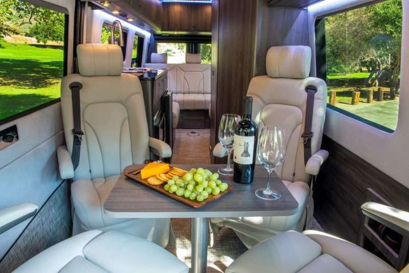 The forward lounge area of the Strada Class B motorhome has four chairs with a removable table. The table is set with a snack board featuring yellow cheese, crackers, and green grapes. There's also a bottle of wine and two wine glasses set out.