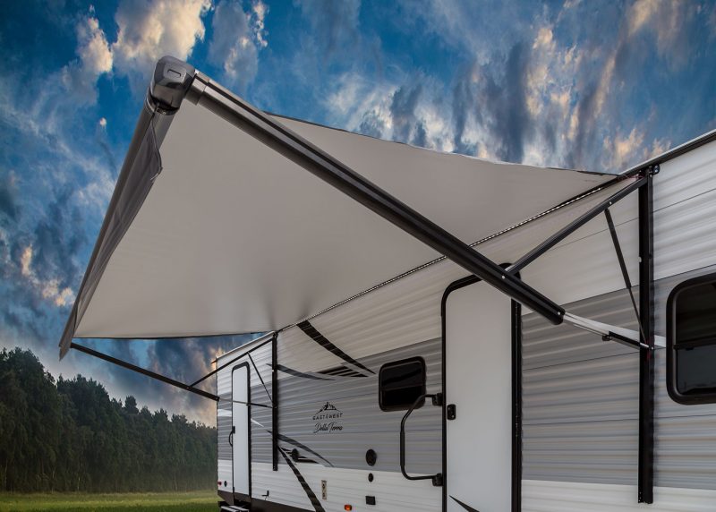 A Della Terra travel trailer is parked with it's awning extended. Clouds are scattered across the blue sky and a row of trees are behind the camper.