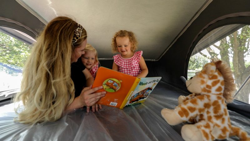 A mother reads to her two daughters while they lay on a bunk bed inside an RV.