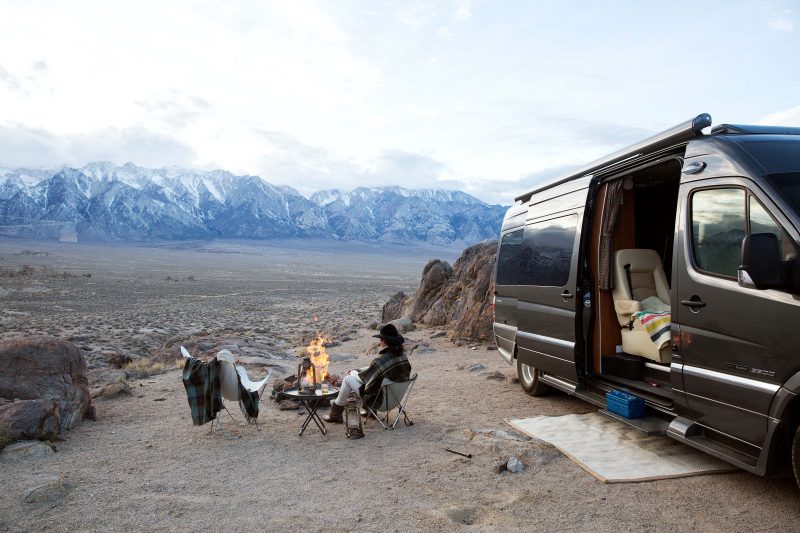 A woman sitting around a campfire near her Class B motorhome that is overlooking a mountain range in the distance.