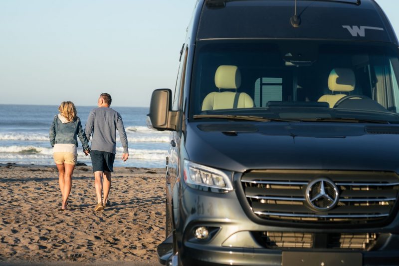 A man and woman hold hands as they walk towards the water on a beach where their Class B RV is parked.