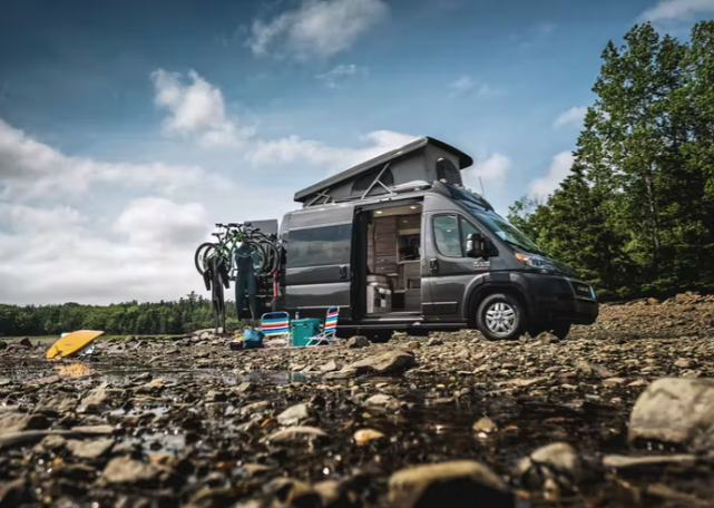 A Thor Scope camper van is parked on a rocky site near water and mature trees.